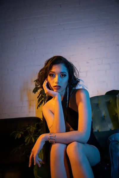 Fashionable asian woman in evening dress looking at camera while sitting on couch in night club — Stock Photo