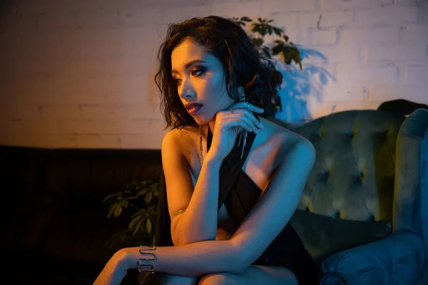 Elegante junge Asiatin im Kleid sitzt auf der Couch in einem modernen Nachtclub mit Beleuchtung — Stockfoto