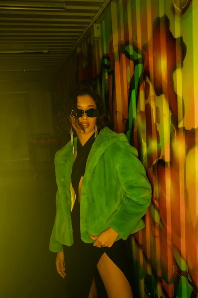 Belle femme asiatique en lunettes de soleil et veste en fausse fourrure debout près de graffitis dans la boîte de nuit — Photo de stock