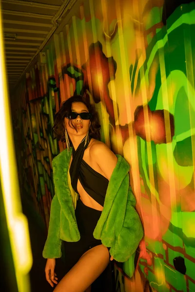 Sexy mujer asiática en gafas de sol y chaqueta de piel falsa de pie cerca de graffiti en el club nocturno - foto de stock