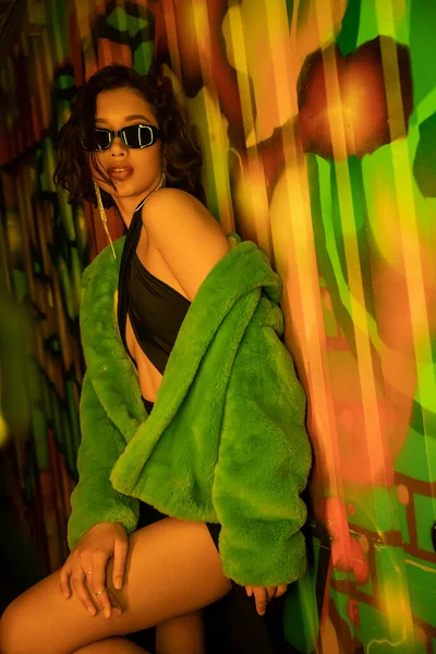 Fashionable young asian woman in sunglasses and sexy dress standing near graffiti in night club — Stock Photo