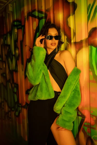 Sexy asiático mulher no falso casaco de pele tocando óculos de sol e de pé perto graffiti no clube noturno — Fotografia de Stock