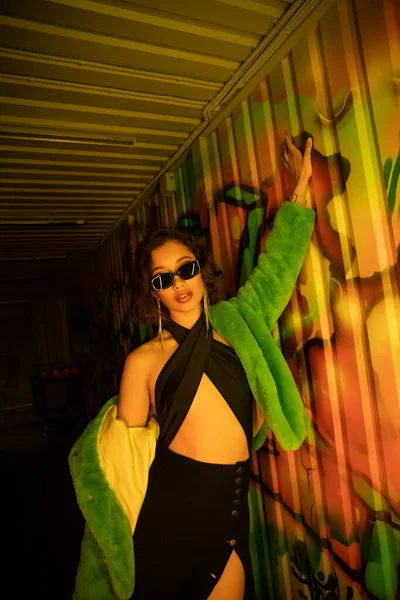 Sexy jovem mulher asiática em óculos de sol e vestido posando perto de graffiti na parede no clube noturno — Fotografia de Stock