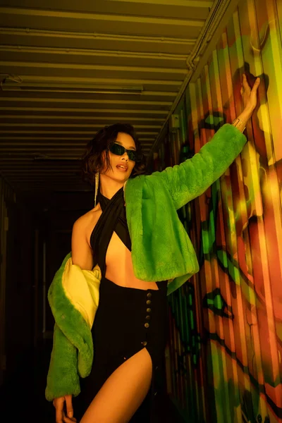 Mulher asiática bonita e sexy em óculos de sol e casaco de pele falso posando perto de grafite na boate — Fotografia de Stock