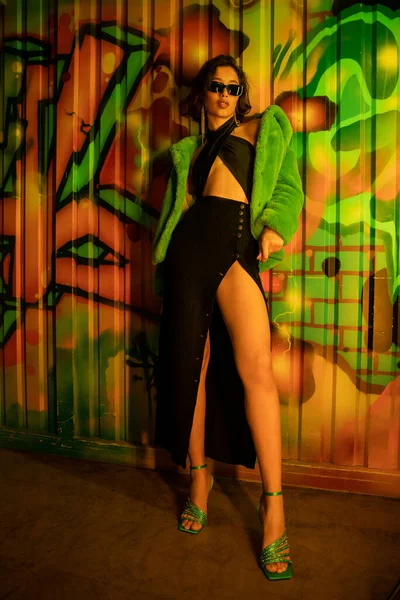 Moda mulher asiática nos saltos, vestido e óculos de sol de pé perto graffiti na parede no clube noturno — Fotografia de Stock