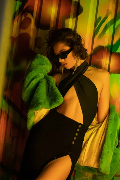 Sexy et élégant asiatique femme en lunettes de soleil et veste en fausse fourrure debout près de graffitis dans la boîte de nuit — Photo de stock