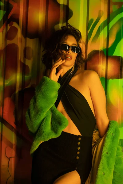 Pretty asian woman in sunglasses and sexy dress standing near graffiti in wall in night club — Stock Photo