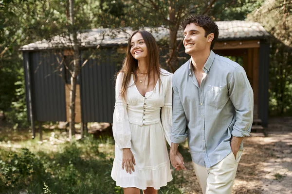 Coppia positiva, uomo e donna felici in prendisole che si tengono per mano, passeggiando vicino alla casa per le vacanze, estate — Foto stock