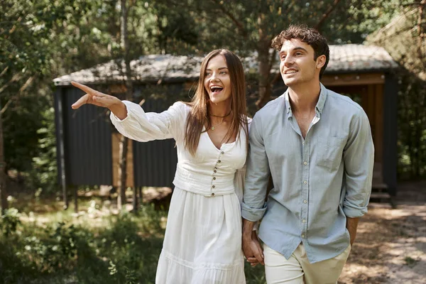 Excited woman in white sundress pointing away, holding hands with man near vacation house, summer — Stock Photo