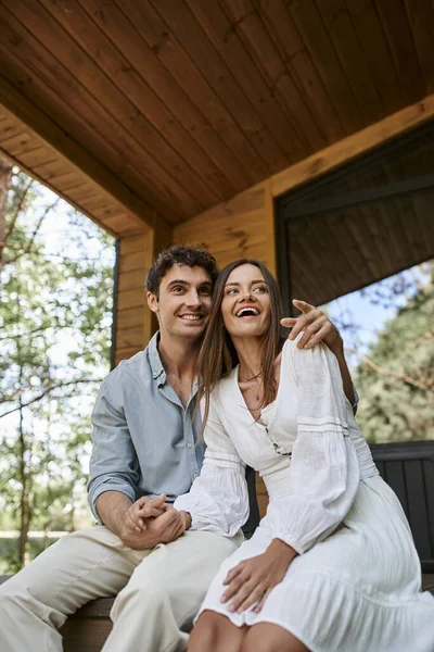 Mann zeigt weg und umarmt fröhliche Frau, während sie auf Veranda sitzt, glückliches Paar, Ferienhaus — Stockfoto