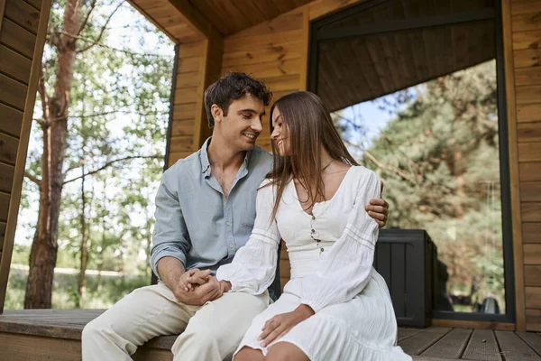 Homme tenant la main avec femme gaie tout en étant assis sur le porche, couple heureux, maison de vacances, romance — Photo de stock
