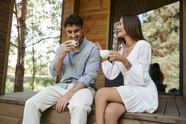 Happy man and woman holding cups of morning coffee, couple laughing on porch of vacation house — Stock Photo
