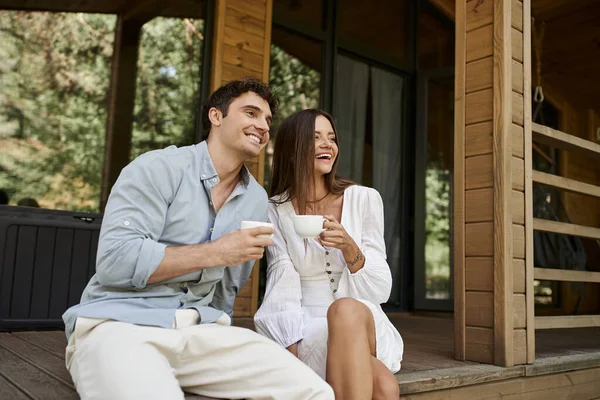 Feliz pareja sosteniendo tazas de café de la mañana, hombre y mujer riendo en el porche de la casa de vacaciones - foto de stock