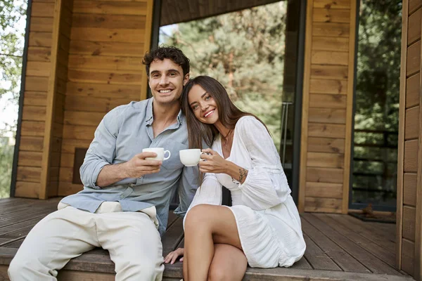 Alegre pareja sosteniendo tazas de café, hombre y mujer riendo en el porche de la casa de vacaciones, mañana - foto de stock