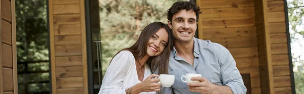 Pancarta, alegre pareja sosteniendo tazas de café, hombre y mujer riendo en el porche de la casa de vacaciones - foto de stock