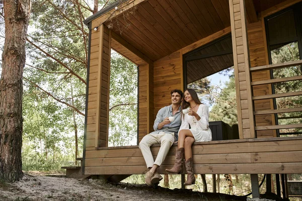 Heureux couple tenant tasses de café, homme et femme souriant sur le porche de la maison de vacances, boisson chaude — Photo de stock