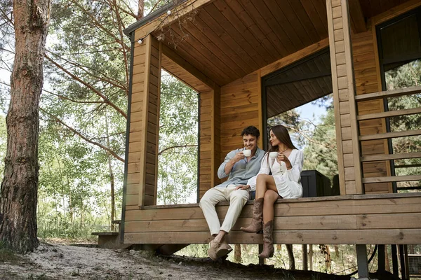 Счастливая пара с чашками кофе, мужчина и женщина улыбаются на крыльце домика отдыха в лесу — стоковое фото