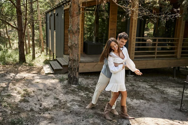 Homme heureux étreignant femme et souriant près de la maison de vacances, forêt, été et romance, couple amoureux — Photo de stock