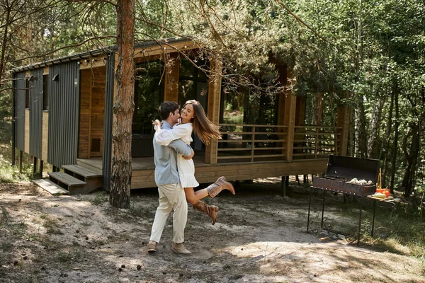 Risa, hombre feliz levantando a mujer alegre con tatuaje, casa de vacaciones en bosque, escapada de verano - foto de stock