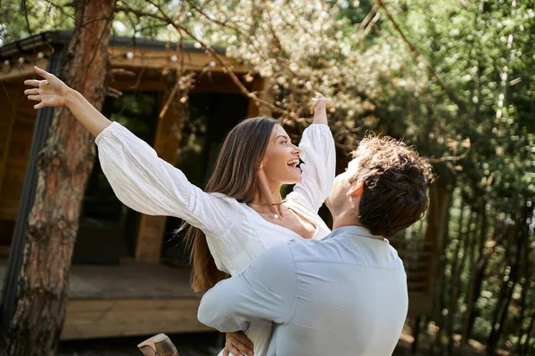 Happy man lifting woman with outstretched hands, vacation house near forest, romance and love — Stock Photo