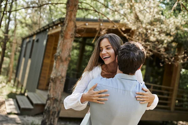 Bel homme étreignant femme heureuse en robe de soleil blanche, maison de vacances près de la forêt, romance et amour — Photo de stock