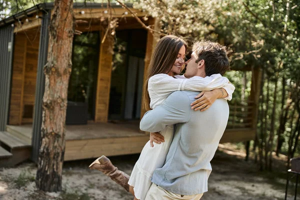 Homem positivo abraçando mulher feliz em sundress branco, casa de férias perto da floresta, romance e amor — Fotografia de Stock