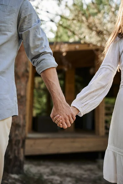 Cropped view, woman in sundress holding hands with man, blurred summer house, romance and love — Stock Photo