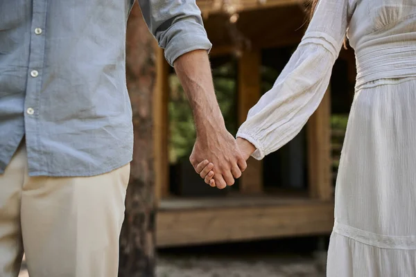 Cropped view, woman in sundress holding hands with man, blurred summer house, love and romance — Stock Photo