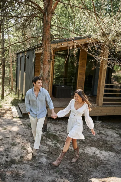 Femme en robe blanche tenant la main et marchant avec son petit ami, souriant près de la maison d'été, romance — Photo de stock