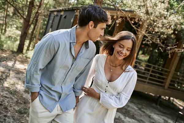 Mujer feliz cogida de la mano con el novio, pareja positiva caminando cerca de la casa de verano en el bosque, el amor - foto de stock