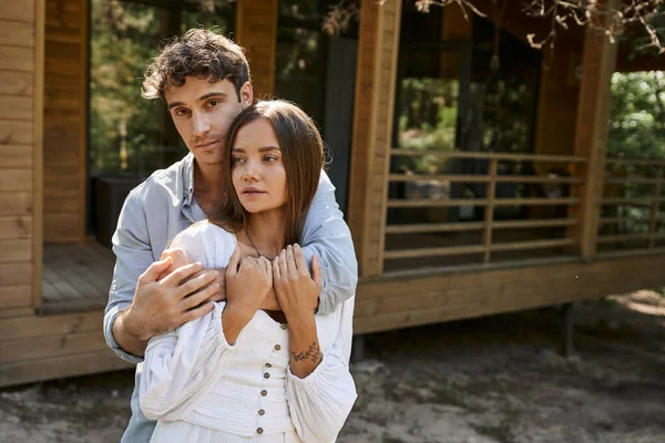 Brünetten Mann umarmt attraktive Freundin in Sundress mit Tätowierung in der Nähe von Ferienhaus im Freien — Stockfoto