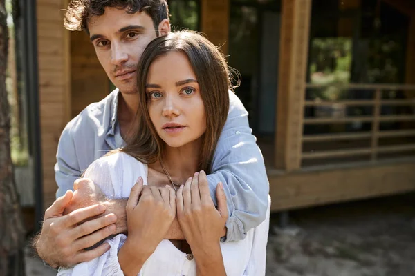 Morena homem abraçando namorada e olhando para a câmera perto de casa de férias borrada ao ar livre — Fotografia de Stock