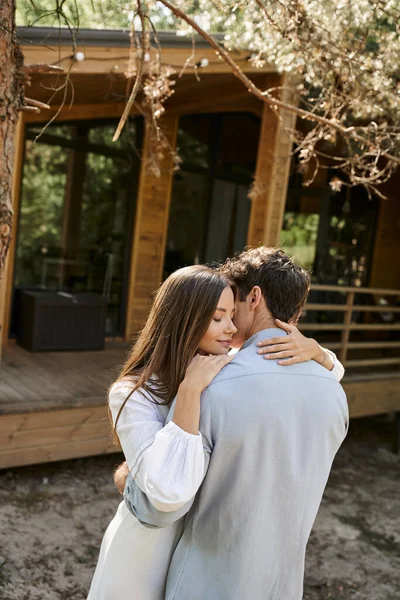 Morena mujer en vestido de sol abrazando novio cerca borrosa casa de verano en el fondo al aire libre — Stock Photo