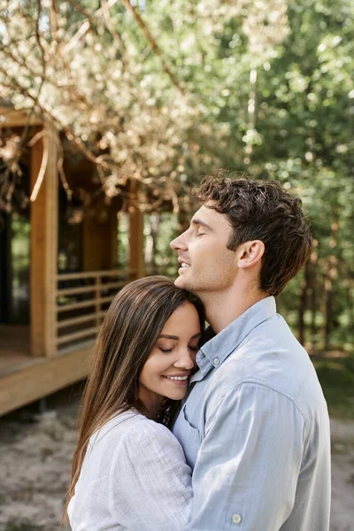 Sonriente pareja con los ojos cerrados abrazando y de pie cerca de la casa de vacaciones en el fondo al aire libre - foto de stock