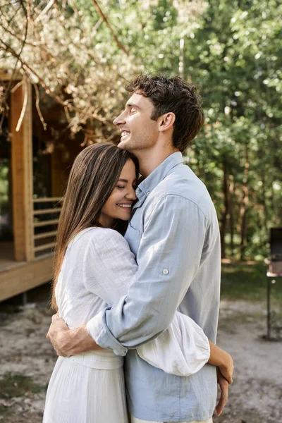 Pareja morena positiva con los ojos cerrados abrazando y de pie cerca de la casa de verano en el fondo - foto de stock