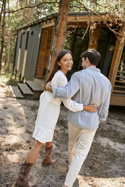Trendy and smiling woman in sundress hugging boyfriend and looking at camera near vacation house — Stock Photo