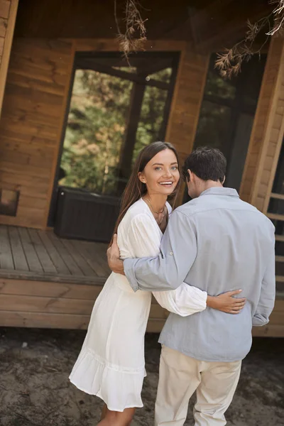 Gioioso giovane donna in sundress abbracciare fidanzato e guardando la fotocamera vicino casa vacanza all'aperto — Foto stock
