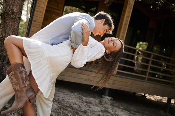 Улыбающийся брюнетка мужчина обнимает подружку в сарафане с закрытыми глазами рядом с домом отдыха на открытом воздухе — стоковое фото