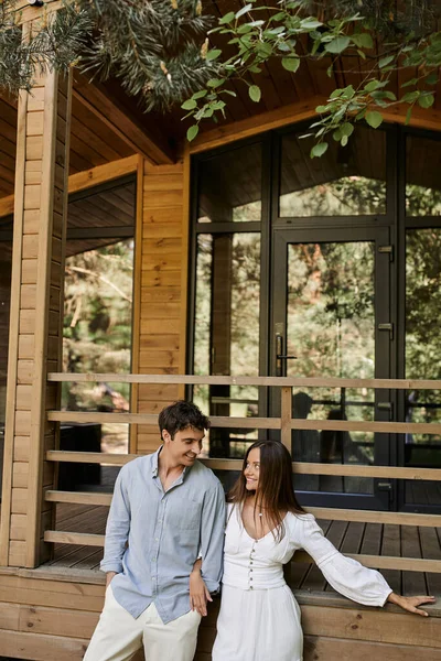 Sonriente y elegante pareja romántica mirándose mientras están de pie cerca de la casa de vacaciones - foto de stock