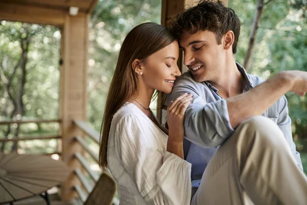 Femme brune souriante en robe de soleil étreignant petit ami avec les yeux fermés près de la maison de vacances en plein air — Photo de stock