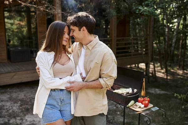 Alegre pareja romántica abrazándose de pie cerca de la barbacoa y la casa de vacaciones durante el picnic al aire libre — Stock Photo