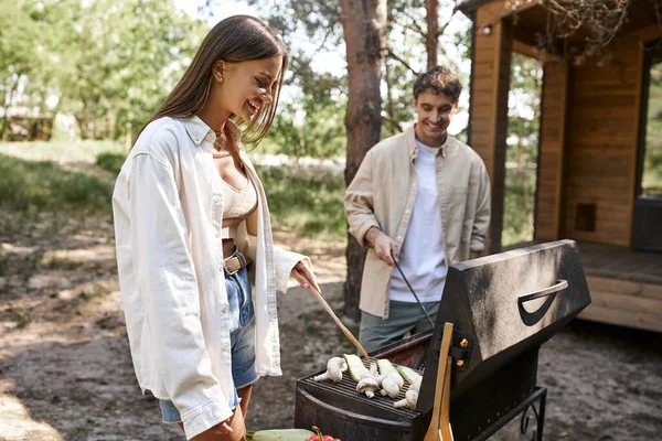 Улыбающаяся женщина готовит овощи на гриле рядом с размытым парнем и домиком для отдыха во время пикника — стоковое фото