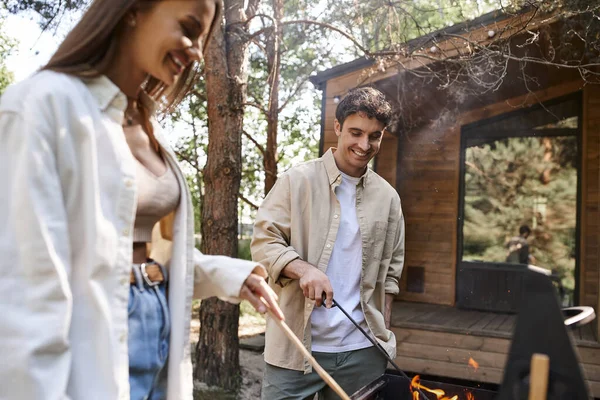 Улыбающийся мужчина готовит на барбекю рядом с размытой подругой и летним домиком на заднем плане — стоковое фото