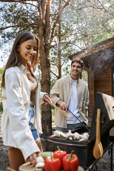 Un homme souriant cuisine sur un barbecue près d'une copine floue avec des légumes et une maison de vacances en plein air — Photo de stock