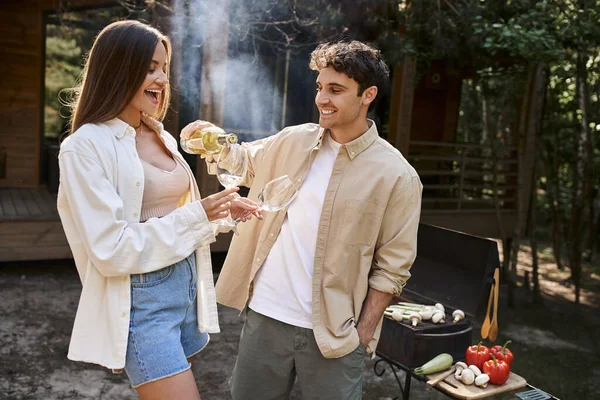 Homme souriant versant du vin près de la petite amie, barbecue flou et maison de vacances à l'arrière-plan — Photo de stock