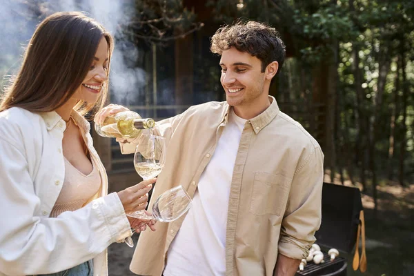 Sorrindo homem derramando vinho perto da namorada e bbq borrado com casa de férias ay fundo ao ar livre — Fotografia de Stock