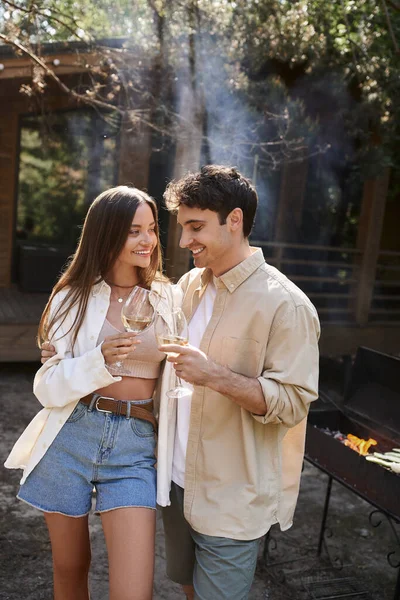 Couple souriant tenant du vin près du barbecue flou et de la maison de vacances en arrière-plan à l'extérieur — Photo de stock