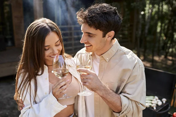 Homme joyeux étreignant petite amie avec du vin près du barbecue et maison de vacances à l'arrière-plan — Photo de stock