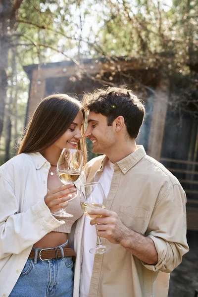 Усміхнена брюнетка пара тримає вино і стоїть з закритими очима біля будинку відпочинку на відкритому повітрі — стокове фото