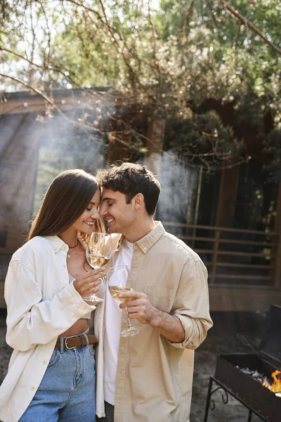 Lächelndes brünettes Paar in lässiger Kleidung mit Wein beim Picknick in der Nähe von Ferienhaus im Freien — Stockfoto
